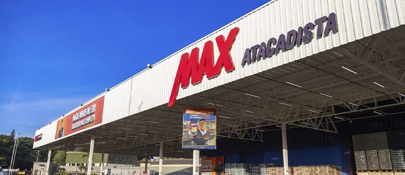 Fachada do Max Atacadista em Sarandi, com o letreiro vermelho da loja sobre a estrutura branca e o céu azul ao fundo