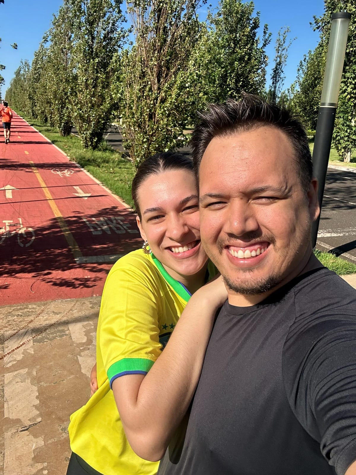 Casal sorrindo na ciclovia arborizada de Maringá, com a pista vermelha de caminhada e corredores ao fundo