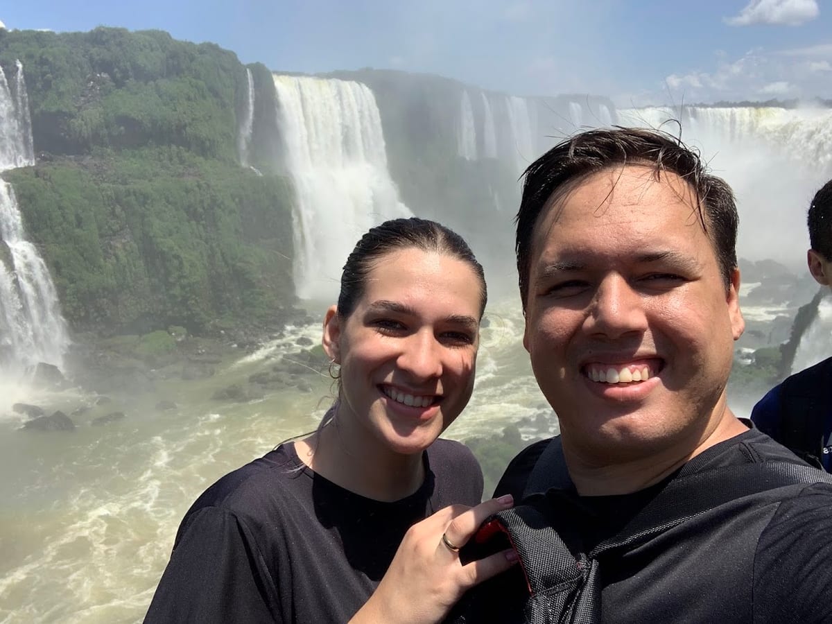 Casal sorrindo e encharcado na passarela das Cataratas do Iguaçu, com as quedas e o cânion do rio ao fundo
