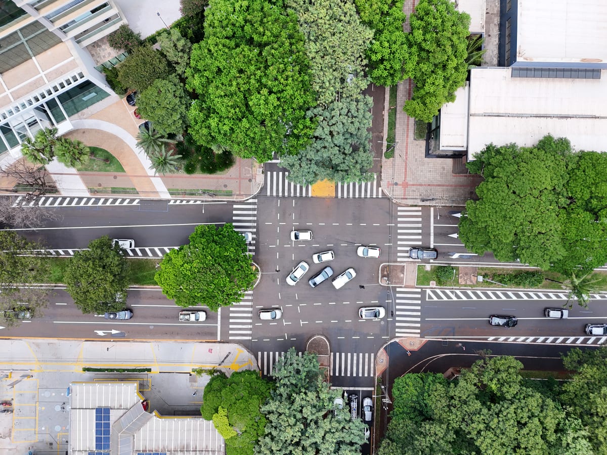 Vista aérea de um cruzamento de Maringá, com faixas de pedestres nos quatro lados, carros parados no meio do cruzamento e árvores nas calçadas